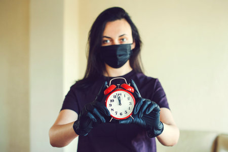 Young woman wearing protective face mask and disposable glove. Woman holds alarm clock. Coronavirus...
