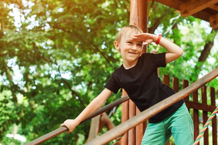 Boy playing in a tree house and looking into the distance. Cute kid playing games at park. Summer vacations, travel and leisure. Happy boy having fun at summer park. Adventure park for kids.の写真素材