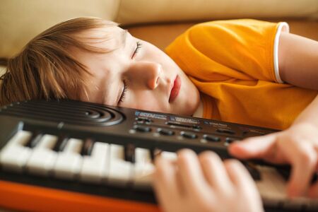 Cute child tired of learning to play the synthesizer. Little dreamer want to be a musician. Tired boy sleeping with synthesizer on the sofa. Kids hobbies and leisure. Future profession.の写真素材