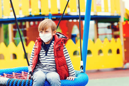 Sad boy in face mask playing outdoors. Bored child at empty playground outside. Coronavirus quarantine. Child wearing safety mask. Coronavirus prevention. Sad kid riding on a swing. Real life 2020.の写真素材
