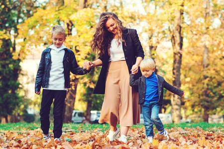 Happy mother and kids having fun outdoors. Fashionable family walking in autumn park. Autumn fashion, lifestyle. Happy children with young mother playing with autumn leaves.の写真素材