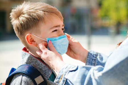 Mother puts a safety mask on her son's face. Schoolboy is ready go to school. Cute boy with a backpack outdoors. Back to school concept. Medical mask to prevent coronavirus. Coronavirus quarantineの写真素材
