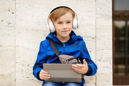 Cute young boy listening to music on tablet outdoors. Kid using wireless headphone. Stylish boy walking the street and enjoying music. People, technology and lifestyle. Handsome kid outside. Fashion.の写真素材