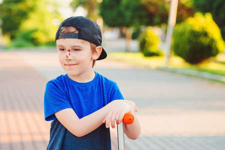 Cute boy learning to ride scooter. Little child after crashing. Active outdoors game, sport. Falling scooter accident. Abrasions on the face. Childhood.の写真素材