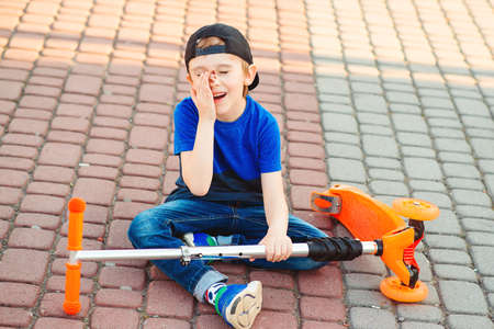 Cute boy learning to ride scooter. Little child after crashing. Active outdoors game, sport. Falling scooter accident. Abrasions on the face. Childhood.の写真素材