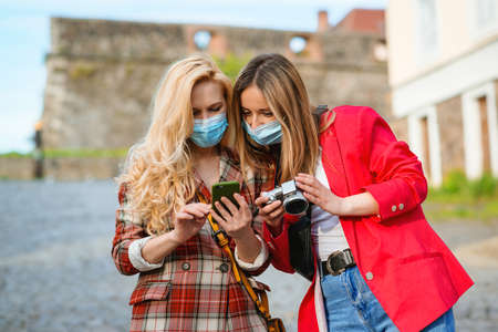 Happy girls traveling together in Europe during pandemic. Stylish women walking in street. Girls taking photos on digital camera. Friends having fun at summer vacations. Female wearing face masks.の写真素材