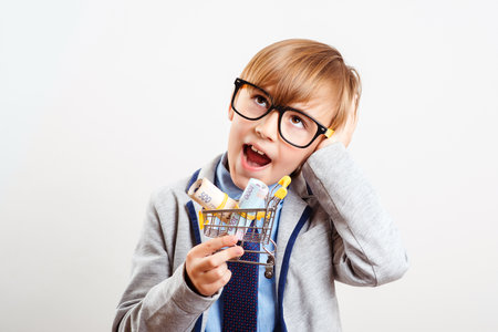 Cute boy with shopping cart isolated on white. Online shop. Little kid holding shopping trolley with money. Child is ready for shopping. Holidays, sales and discounts. Nerd child in glasses and suit.の写真素材