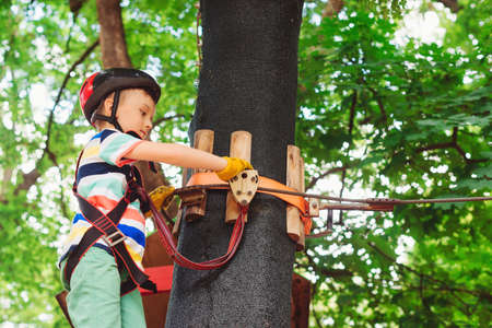 Cute boy in the rope park pass obstacles. Child climb the rope road. Boy wears safety helmet and equipment. Summer camp. Kid is ready to start climbing obstacles.の写真素材