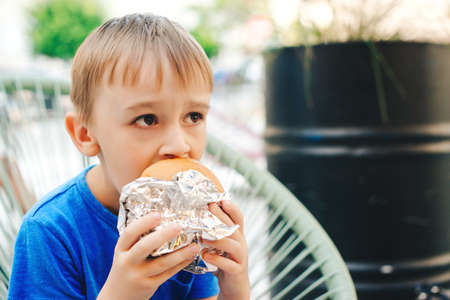 Hungry kid eating a burger at outdoors cafe. Cute child eating fast food. Childhood, unhealthy food concept. Junky food. Lifestyle and peopleの写真素材