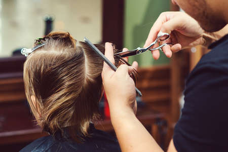 Cute little boy getting haircut by hairdresser at the barbershop. Barber man doing kid the hairstyle. Hairdresser with scissors. Barber shop. Childhood. New hairstyle for young boy.の写真素材