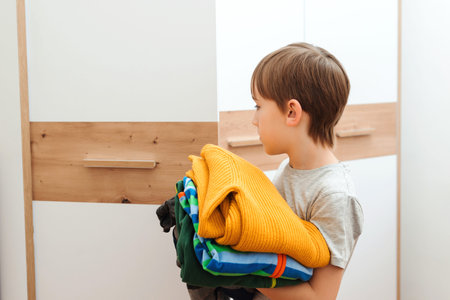 The boy puts things in order in the closet. A stack of colorful clothes. Kid organizing clothes in...