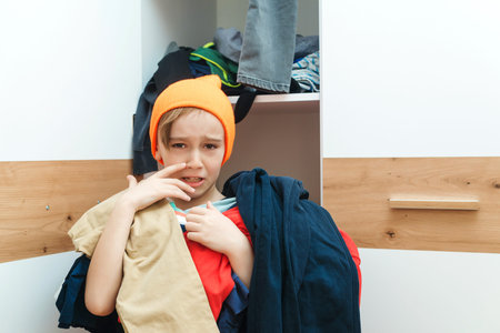 Boy holding pile of dirty clothes. Untidy clutter clothing closet. Messy home kid's room. Home chores housework. Mess in the wardrobe. Many scattered clothes in children bedroom.の写真素材