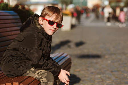 Handsome boy sitting on the bench in the city. Preteen boy wearing black jacket and trendy sunglasses. Stylish street style boys outfit. Fashion, lifestyle and people. Young boy wearing sunglasses.の写真素材