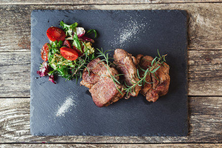Grilled beef steaks on black stone slate, top view. Tasty juicy grilled steak and salad with tomatoes. Main course beef dinner. Dinner with steak beef and salad. Big stake meat on slate.の写真素材