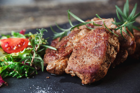 Grilled beef steaks on black stone slate. Tasty juicy grilled steak and salad with tomatoes. Main...