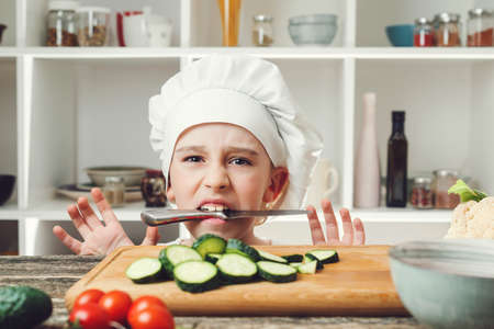 Angry chef with knife in mouth. Child want to be a professional chef. Boy chef cutting vegetables for salad. Cook with knife. Cute child wearing chef hat and uniform.の写真素材