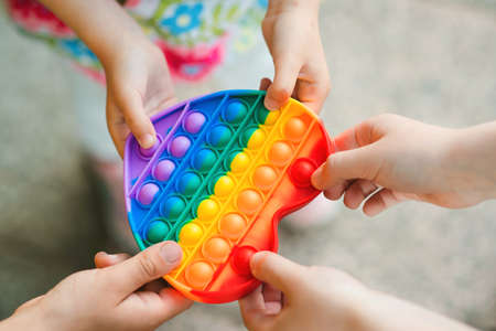Group of kids holding popular toy, top view. Happy kids having fun outdoors. Modern anti stress toy in heart shape. Friends on a walk with silicon bubble toys. Children hands with rainbowの写真素材