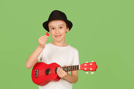 Cute boy plays the ukulele. Happy kid enjoying the music. Student learning to play ukuleles. Fashionable boy in summer hat isolated over green background.の写真素材