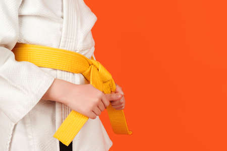 Kid in karate kimono holding his yellow belt, close up. Boy practicing karate on color background, copy space. Kid sport concept. Healthy sporty childhood and lifestyle. Martial arts, school and training.の写真素材