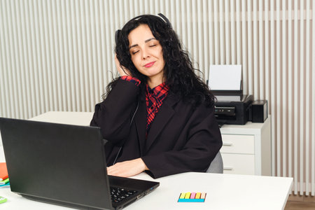 Businesswoman listening to music in headphones during work break. Young smiling employee wearing...