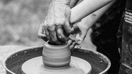 Potter teaching child to make ceramic pot on the pottery wheel. Pottery workshop outside. Potters and child hands. Family working on pottery wheel. Dad and child making ceramic pot together.の写真素材