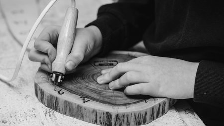 Boy hands burn out numbers with soldering iron on wooden disc. Kid makes wooden clock in the...