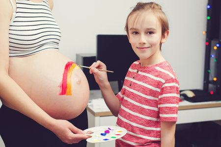 Cute boy drawing rainbow on pregnant belly his mother. Baby birth expecting time and belly painting. Pregnant mom and her son having fun together at home. Family, healthy pregnancy.の写真素材
