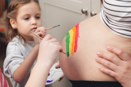 Happy little girl drawing rainbow on pregnant belly her mother. Baby birth expecting time and belly painting. Pregnant mom and child having fun together at home.の写真素材