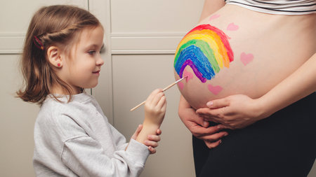 Sweet little daugter painting pregnant belly her mother. Baby birth expecting time and belly painting. Pregnant mom and her child having fun together at home. Family, healthy pregnancy and baby birth.の写真素材