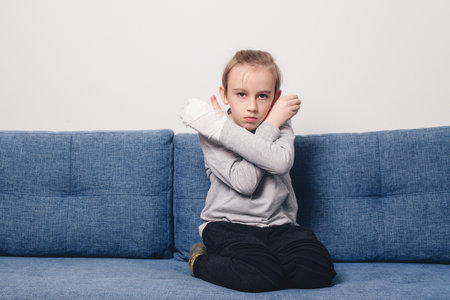 Sad boy with broken hand at home. Kid with broken arm and gypsum sitting on the sofa. Injury, traumas, accident by kids. Health care, medicine and childhood concept.の写真素材