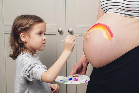 Sweet little daugter painting pregnant belly their mother. Baby birth expecting time and belly painting. Pregnant mom and her child having fun together at home. Family, healthy pregnancy, baby birth.の写真素材