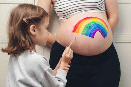 Sweet little daugter painting pregnant belly her mother. Baby birth expecting time and belly painting. Pregnant mom and her child having fun together at home. Family, healthy pregnancy and baby birth.の写真素材