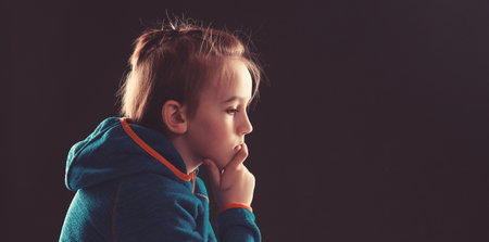 Stressed boy over black background, copy space. Childhood, bulling and family concept. Sad young boy thinking about his problems. Afraid child sitting quietly in the dark. Young boy suffering from anxiety.の写真素材