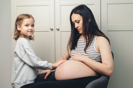 Happy pregnant woman, her child, expectation. Adorable little preteen girl cuddling mother's pregnant belly. Pregnant woman with her little daughter at home. Cute little girl is hugging mom's tummy.の写真素材