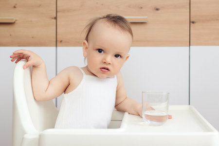 Sweet baby girl holding glass and drinking water. Happy baby sitting in a baby chair in the kitchen. Child drinks water.の写真素材