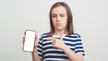 Serious boy holding mobile phone with white blank screen in hand. Gadget with empty free space for mock up. Young student boy in casual. Boy pointing at empty free space on smartphone.の写真素材