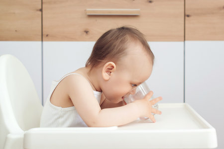Sweet baby girl holding glass and drinking water. Happy baby sitting in a baby chair in the kitchen. Child drinks water.の写真素材
