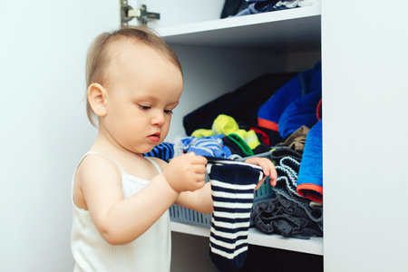 Baby girl takes clothes from wardrobe herself. Little daughter playing with garments at home. Toddler chooses an apparel. Real life with toddler at home.の写真素材