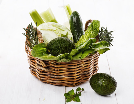 Fresh organic green vegetables and herbs in a woven basket on white wooden backgroundの写真素材