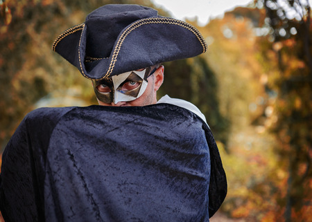 Mystery man in masquerade mask, triangle hat and black velvet cape hiding his lower face with his hand. Autumn (fall) forest background, shallow depth of fieldの写真素材