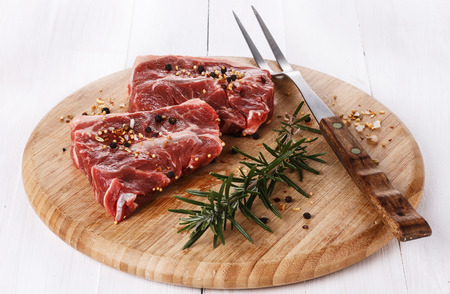 Two pieces of red meat, rosemary, spices  and fork on a round cutting board over white wooden . Copy spaceの写真素材
