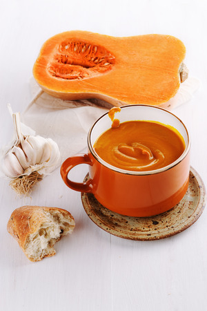 Bowl of creamy pumpkin soup with clove of garlic and bread in a rustic ceramic plate on a white wooden backgroundの写真素材
