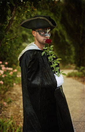 Retro image of romantic mystery man in black mask and velvet cape holding a red rose. Selective...