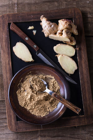 Top view image of ground ginger in a ceramic plate and fresh ginger root cut on the pieces on black board over rustic wooden backgroundの写真素材