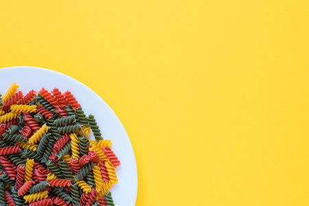 Red green and yellow dry raw fusilli pasta on a plate with yellow background and copy space.
