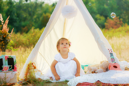 A girl in a white dress sits in a white tent on a background of green bushes. The child sat on his haunches.の写真素材