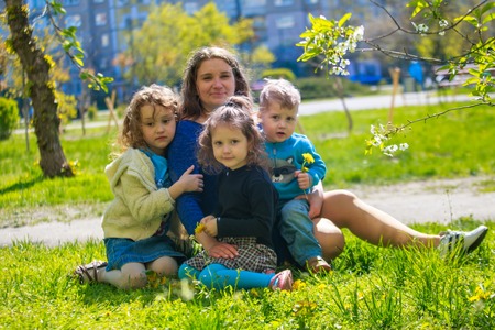 Little girl hugs mother sitting on the grass. Child, playing, threw his mother on grass. Family in spring walks on street.の写真素材