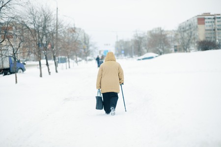 An elderly woman with cane is walking along a snow-covered pavement.の写真素材