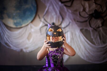 girl hid her face behind the pumpkin at Halloween. girl in the witch costume frightens. child is celebrating at All Saints Day. Scary spider on the wall. Cut black pumpkin. Decor for Halloween.の写真素材