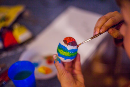 child decorates an Easter egg in the colors of the rainbow. Boy holds an egg and paints it with brush. Preparing for celebration of Easter.の写真素材